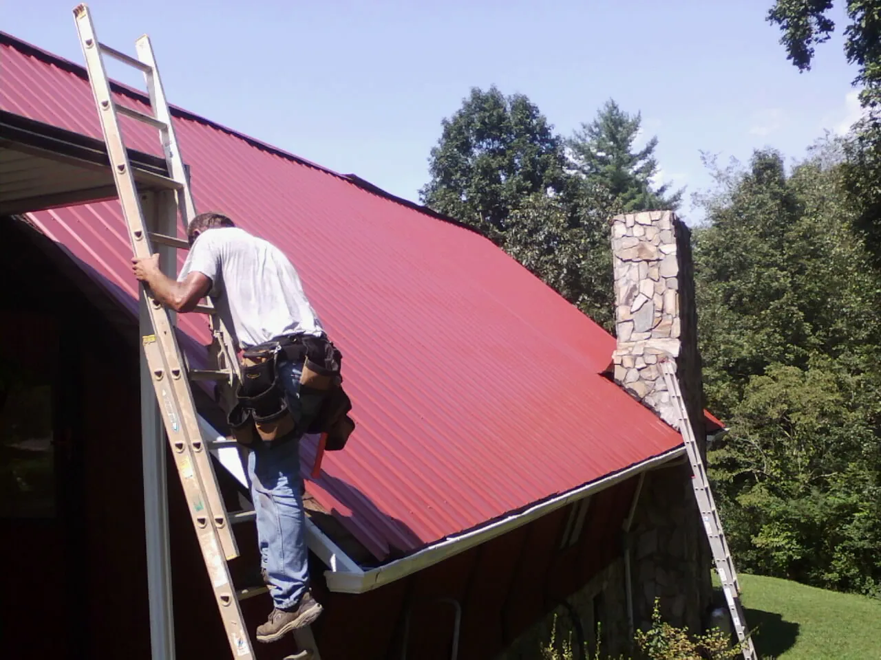 Roofer on a ladder beside a red metal roof and stone chimney — Soffit & Fascia Repair by Roofing Brothers in Linden